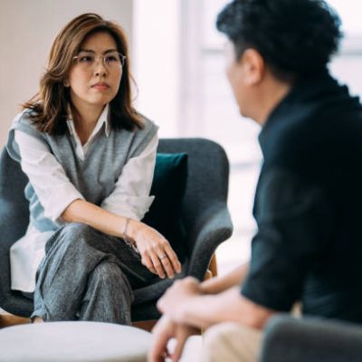 An Asian female professional psychologist and a mature man are sitting on a cozy sofa, discussing the problems he is facing during a therapy session at a comfortable office.